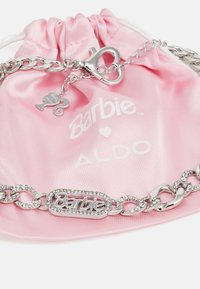 Sølvsmykke armbånd med "Barbie"-logo omgitt av rhinestone, plassert på en rosa sateng pose med snøring merket med "Barbie" og "ALDO."