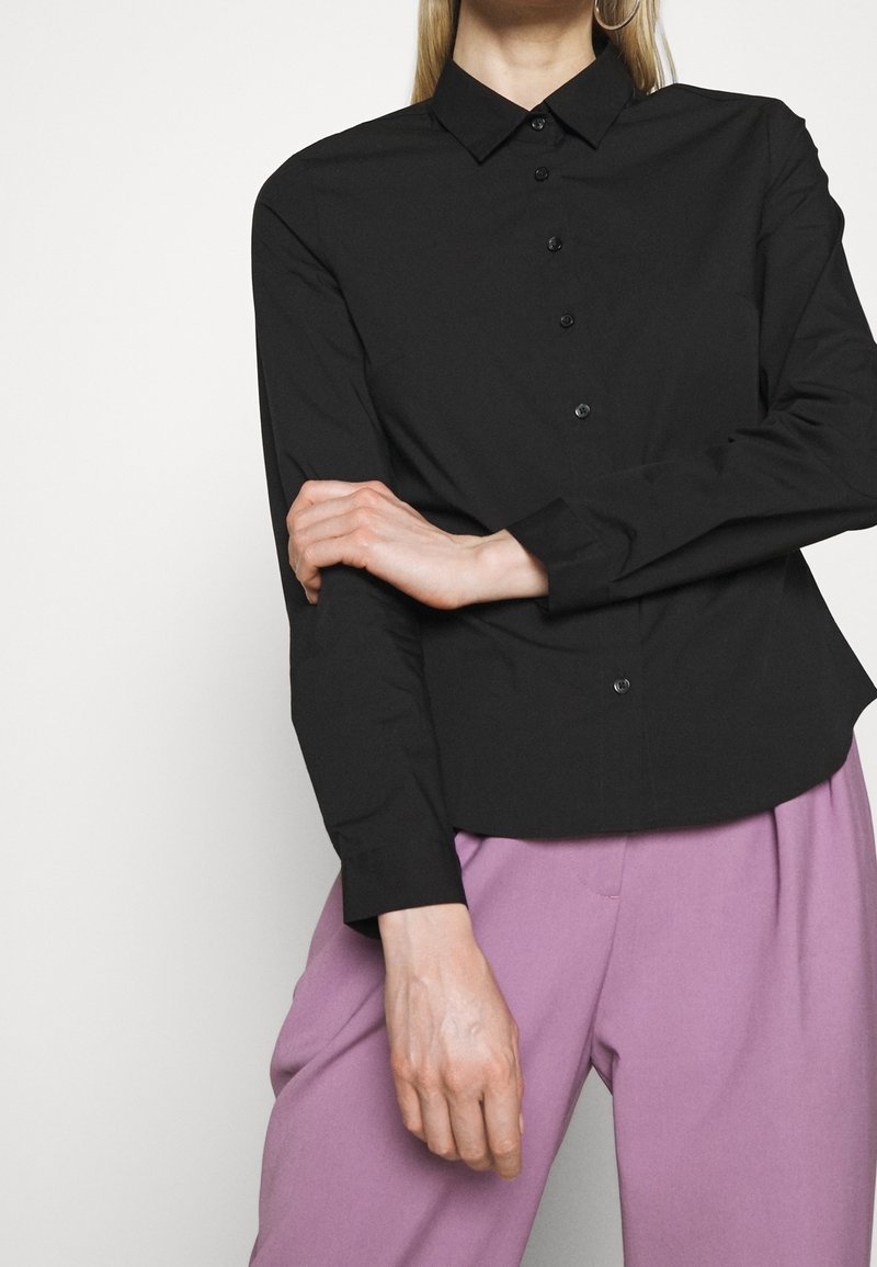 Femme portant une chemise noire à manches longues boutonnée avec col et un pantalon violet clair, les bras croisés à la taille devant un fond uni.