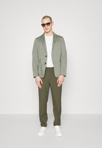 Hellgrünes Blazer mit zwei Knöpfen, weißem T-Shirt, olivgrünen Hosen und weißen Sneakers. Texturiertes Gewebe mit schlanker Passform.