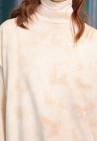 Žena v béžovém tričku s žíháním a rolákem, na hrudi s bílým logem Nike, s volně spadajícími hnědými vlasy.