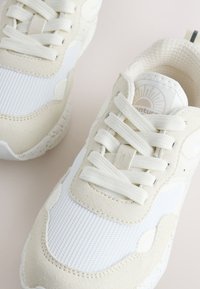 Baskets blanches et beiges avec des empiècements en mesh texturé et en suède, des lacets blancs épais et des collerettes rembourrées sur une surface claire.