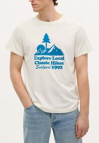 Tricou de culoare crem cu un design grafic albastru, prezentând munți, un copac și text. Mâneci scurte, croi lejer. Țesătură ușoară.