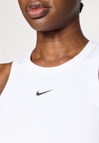 Biały bezrękawnik sportowy wykonany z gładkiego materiału, z czarnym logo Nike na środku klatki piersiowej. Okrągły dekolt, dopasowany krój.