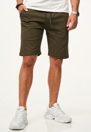 Mann trägt olivgrüne Shorts mit Kordelzug, weiße Turnschuhe, ein weißes Hemd, eine Uhr und ein Armband, steht mit einer Hand in der Tasche.