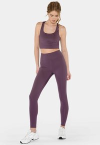 Naine seisab, kandes sobivaid lillakas spordirinnahoidjat ja kõrge vöökohaga retuuse, jalas valged spordijalatsid, vaadates alla ja toetades ühe käega puusa.