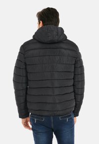 Uomo che indossa un giubbotto imbottito nero a trapunta con cappuccio, in piedi rivolto di spalle, abbinato a jeans blu con tasche posteriori con zip.