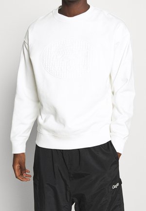 Mann trägt ein weißes Sweatshirt mit erhabenem ovalem Logo und schwarze Shorts mit kleinem weißen Logo am linken Oberschenkel.