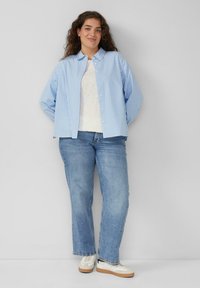  Lichtblauwe en witte gestreepte blouse met lange mouwen, gecombineerd met een witte top en blauwe jeans. Model draagt witte sneakers.