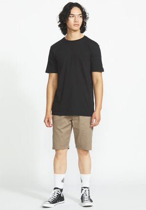 Jeune homme aux cheveux bouclés portant un t-shirt noir, un short beige, des chaussettes blanches et des baskets montantes noires, debout devant un fond uni.