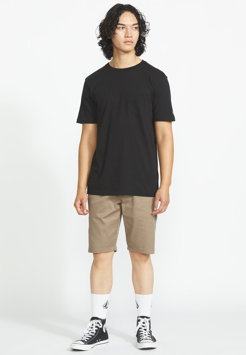 Jeune homme aux cheveux bouclés portant un t-shirt noir, un short beige, des chaussettes blanches et des baskets montantes noires, debout devant un fond uni.