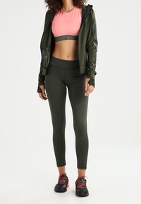 Conjunto deportivo que incluye un crop top rosa, una sudadera con cremallera verde oliva y leggings ajustados también verdes, combinados con zapatos negros y cintas rojas.