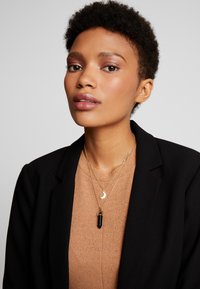 Guldhalskæder lagdelt over en tan ribbet top, der har en halvmånecharms og en lodret sort vedhæng, sat mod en sort blazer.