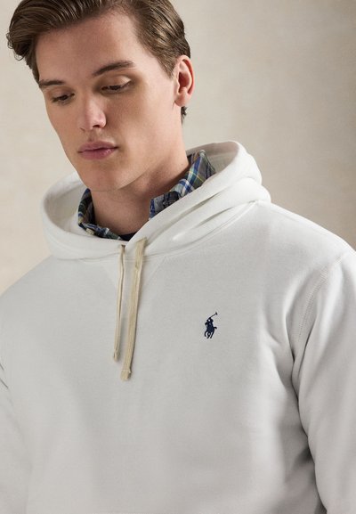 Hvit hettegenser laget av bomull, med en teksturert finish. Har hette med snøring, en liten blå logo, og er lagvis med en rutete skjorte under.