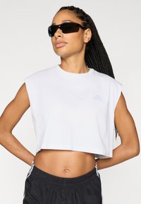 Λευκό αθλητικό τοπ κοντό, με κοντά μανίκια, με ένα ανοιχτό μπλε λογότυπο στην αριστερή πλευρά, σε συνδυασμό με μαύρο σορτς και γυαλιά ηλίου.