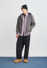 Γκρι φούτερ με φερμουάρ πάνω από μια μοβ και άσπρη ριγέ μπλούζα, συνδυασμένο με μαύρες cargo παντελόνες και καφέ μπότες. Το μοντέλο φοράει μαύρο σκουφάκι.