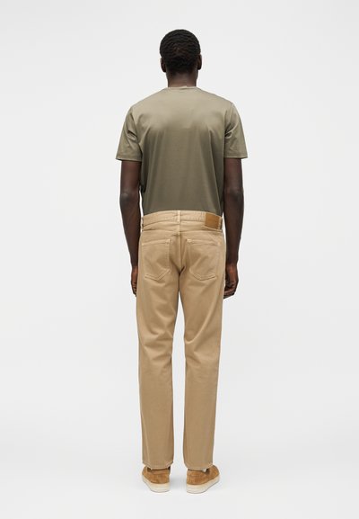 Mees seisab seljaga, seljas oliivroheline t-särk, beežid püksid ja helepruunid jalanõud, taust on ühtlane ja hele.