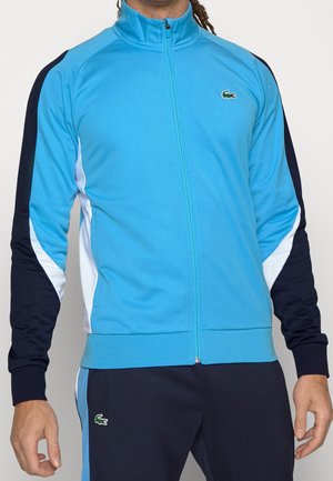 Personne portant une veste zippée bleu clair et bleu marine avec des accents blancs et un pantalon bleu marine assorti, tous deux arborant un petit logo crocodile vert.