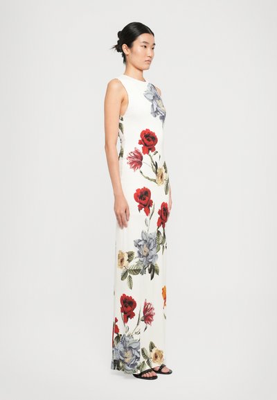 Woman wearing a sleeveless white maxi dress with large red, blue, and yellow floral prints, standing sideways against a plain background.