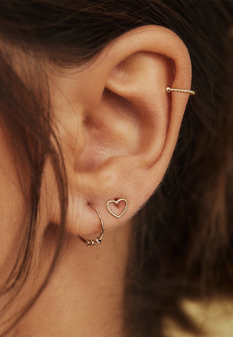Gros plan d'une oreille avec trois boucles d'oreilles en or : manchette torsadée sur le haut de l'oreille, petite puce en forme de cœur et petit anneau avec perles sur le lobe.