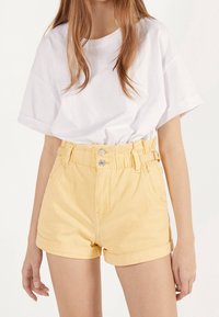 Vit oversized t-shirt och höghöga gula jeansshorts med uppvikta fållar, med en bälte i midjan och silverdetaljer på knappar.