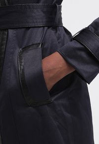 Granatowy płaszcz o teksturowanym wykończeniu, z kieszenią wykończoną czarną skórą i związanym paskiem w talii; widoczne eleganckie, nowoczesne elementy wzornictwa.