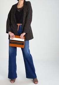 Blazer noir à motifs, top en dentelle noire, jean denim bleu à jambes larges, pochette beige avec rayures blanches et noires, mules à talons grises.