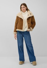 Bruine suède jas met een crèmekleurige nepbontkraag, voorzien van twee grote knopen, gedragen over een witte gebreide trui en blauwe jeans met wijd uitlopende pijpen.