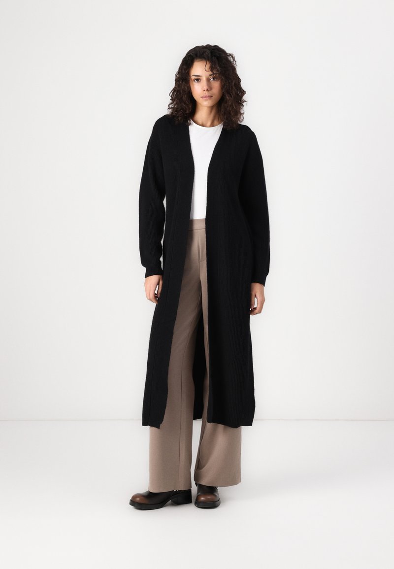 Lang svart strikket cardigan med åpen front, ribbet tekstur og lange ermer, kombinert med beige vide bukser og mørke sko.