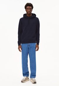 Marineblaue Kapuzenpullover mit einer Vordertasche und verstellbarem Kordelzug; kombiniert mit hellblauen Jeans und grauen Sneakers mit weißen Akzenten.