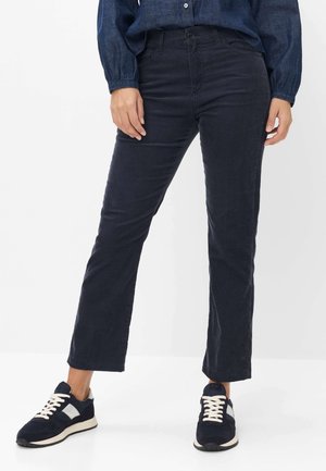 Kvinde iført mørkeblå kordsbukser, marineblå sneakers med hvide snørebånd og en blå denimskjorte, der står mod en hvid baggrund.