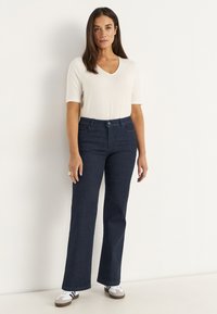 Kvinna med långt brunt hår iklädd en vit t-shirt med kort ärm och V-ringning, mörkblå bootcut jeans och vita sneakers, stående mot en vit vägg.