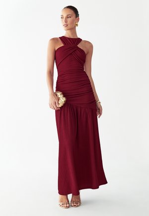 Kobieta w bordowej, marszczonej wieczorowej sukni bez rękawów, trzymająca złotą kopertówkę, z złotymi kolczykami, bransoletką i sandałkami na obcasie.