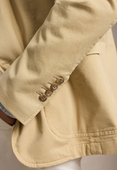 Manche de blazer beige avec quatre boutons marron reposant sur la poche de hanche d'une veste beige assortie portée par une personne.