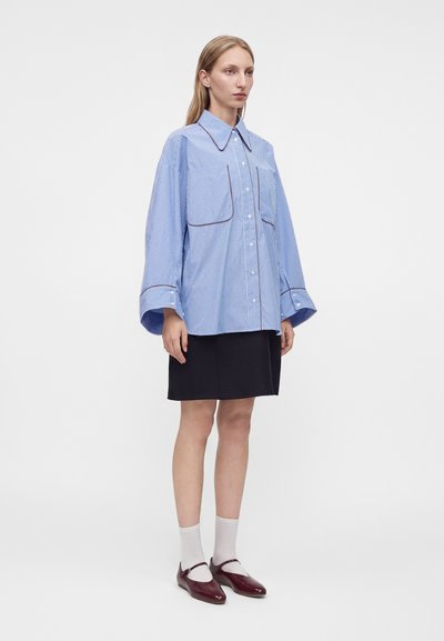 Jeune femme portant une chemise oversize rayée bleue et blanche, une jupe noire, des chaussettes blanches et des chaussures Mary Jane rouge foncé, debout sur fond blanc.