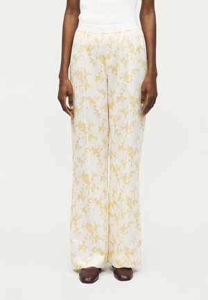 Άτομο που φοράει λευκό παντελόνι με φαρδύ πόδι με ανοιχτό κίτρινο λουλουδάτο μοτίβο και καφέ παπούτσια με τετράγωνη μύτη, στέκεται μπροστά σε απλό φόντο.