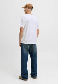 Camiseta blanca de algodón combinada con pantalones vaqueros de denim azul de corte holgado. El modelo lleva una gorra marrón, mostrando una silueta casual.