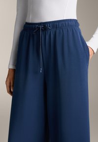 Temno modri široki hlače z elastičnim pasom in podrobnostjo v obliki vrvice. Narejene so iz gladkega materiala in imajo stranske žepke.