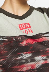 Krátký rukáv sportovního trička s mátově zeleným vrškem, tmavohnědými akcenty a vícebarevným abstraktním vzorem na dolní části. Obsahuje síťovinovou texturu.