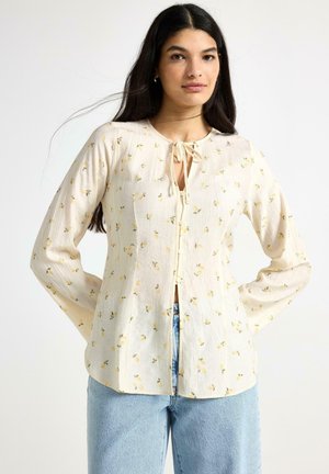 Ung kvinde med langt mørkt hår iført en cremefarvet bluse med små gule blomsterprint og lyseblå jeans, stående med hænderne bag ryggen.