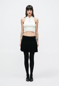 Top corto blanco de canalé con cuello, falda negra acampanada, medias negras y zapatos negros. La modelo está de pie, con fondo minimalista.