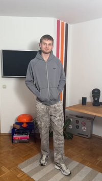 Γκρι φούτερ με φερμουάρ και λογότυπο στο στήθος, συνδυασμένο με ανοιχτόχρωμο παντελόνι cargo με σχέδιο και αθλητικά παπούτσια. Εσωτερικός χώρος με τηλεόραση και διακόσμηση.