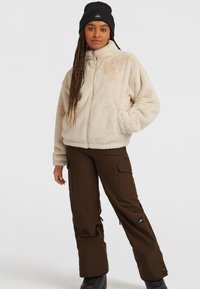 Fuzzy beige zip-up jack met hoge kraag, gecombineerd met bruine cargobroek met zijzakken en zwarte laarzen. Zwarte beanie op het hoofd.