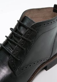 Botín negro de cuero hasta el tobillo con detalles de brogue, puntera redonda y cierre de cordones. Presenta una superficie lisa y una suela marrón de contraste.