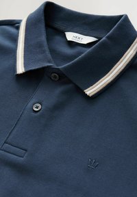 Mørkeblå polo-shirt med tekstureret stof, en to-knaps placket og en kontrasterende stribet krave i creme. Har et broderet logo på brystet.