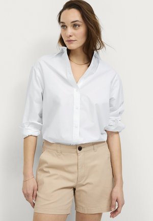 Kvinde iført en hvid skjorte med knapper og opkørte ærmer samt beige shorts, stående mod en ensartet, lys baggrund.