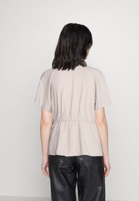 Blusa de un beige claro con mangas cortas, detalle de cintura fruncida y textura suave, combinada con pantalones negros, vista desde la espalda.