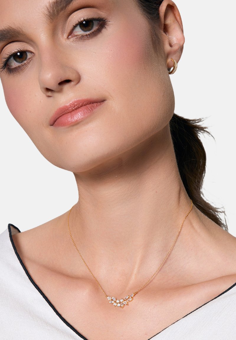 Złoty naszyjnik łańcuszkowy o zakrzywionym wzorze, zdobiony małymi, przezroczystymi kamieniami ułożonymi w wzory kwiatowe. Noszony na nagiej skórze.