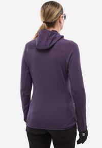 Femme portant une veste à capuche violette, des gants noirs et un pantalon noir, se tenant de dos contre un fond blanc uni.