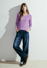 Fioletowa bluzka z długim rękawem i dekoltem w kształcie V, połączona z szerokimi niebieskimi dżinsami. Model nosi czarne sneakersy z brązową podeszwą.