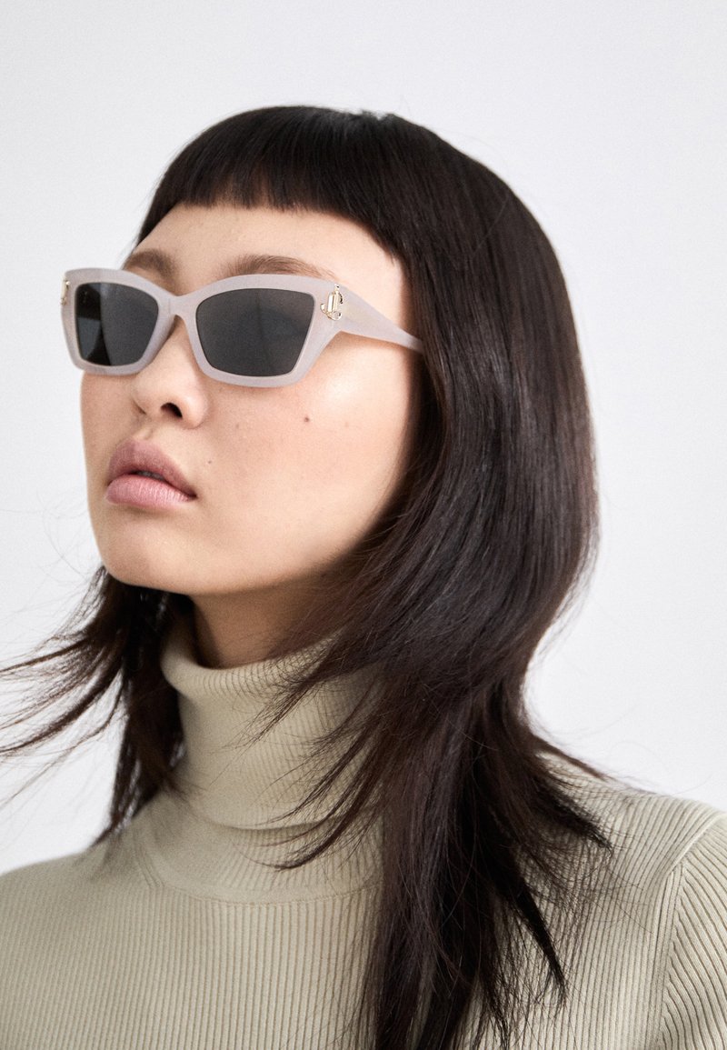 Frosted white cat-eye sunglasses with dark lenses and gold accents. Model wears a ribbed cream turtleneck. Hair styled straight and sleek.